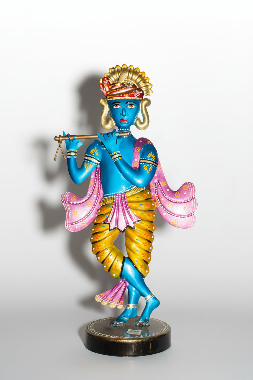A metal table decor depicting Lord Krishna in a blue and gold colored attire, holding a flute, standing on a round base.