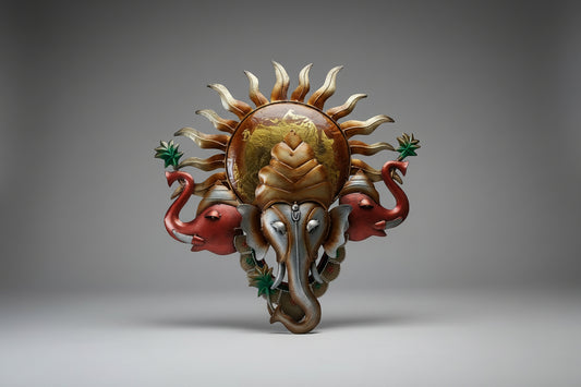 A metal idol of Lord Ganesh with a sun pattern and two elephant hands. The idol is predominantly gold with details in black and green.
