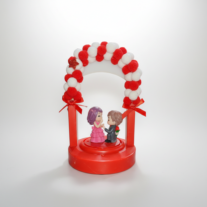 A decorative showpiece featuring a couple figurine standing under a red and white arch with ribbons and pom-poms.